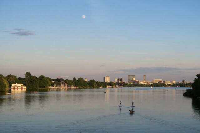 Ruhiger Fluss mit Stadtansicht, Sportler auf dem Wasser und Vollmond im Hintergrund.