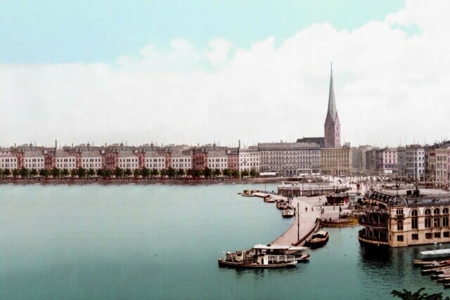 Historische Ansicht eines Hafens mit Gebäuden und Booten, umgeben von Wasser und Himmel.