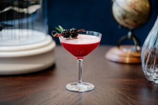 Hotel Atlantic Eleganter Cocktail in einem Glas, rot mit Beeren und Rosmarin, auf einem Holztisch.