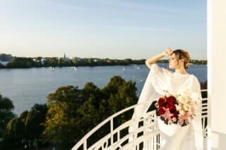 Hotel Atlantic Braut in eleganter Robe mit Blumenstrauß, Blick auf den See und die Stadt im Hintergrund.