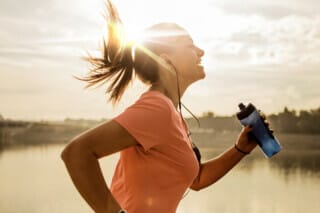 Runner with headphones and water bottle, jogging by the water at sunset.