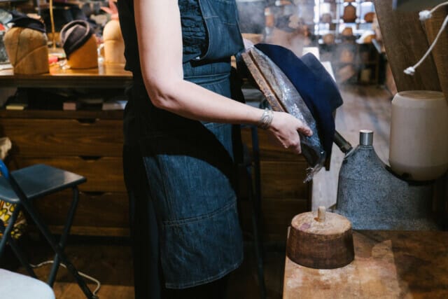 Handwerkerin in einer Hutwerkstatt, die einen Hut mit Dampf bearbeitet. Holzformen im Hintergrund.