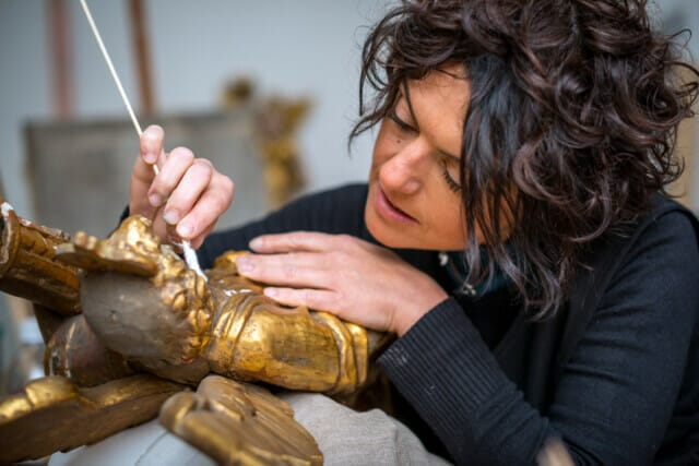 Frau reinigt sorgfältig eine vergoldete Statue mit einem Wattestäbchen.