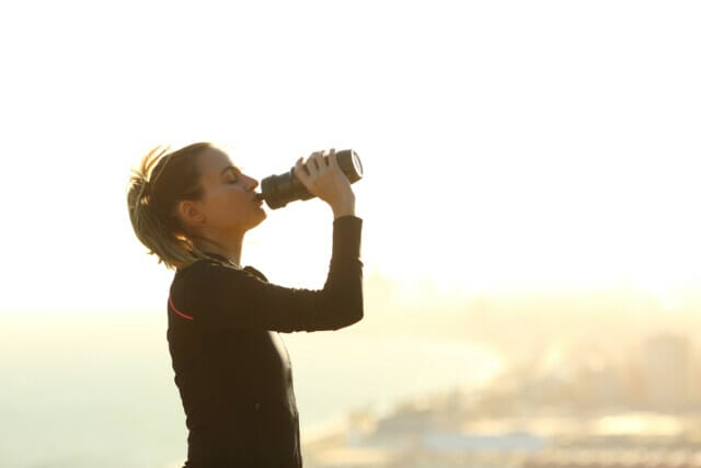 Side view portrait of a runner woman drinking water from bottle hydrating after exercise in a city outskirts