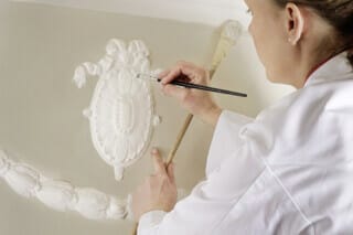 Hotel Atlantic Craftswoman paints filigree stucco decorations on the ceiling in a historic room.
