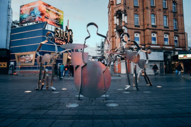 Skulpturen von Musikern in einer Stadtstraße mit buntem Hintergrund und Menschenmenge.