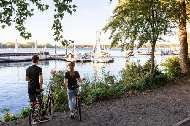 Paar mit Fahrrädern am Seeufer, Blick auf Segelboote und eine malerische Landschaft.