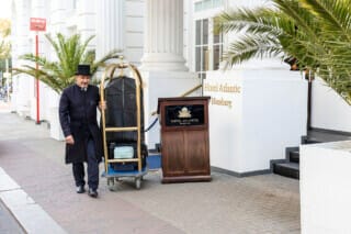 Porter with luggage trolley in front of the Hotel Atlantic in Hamburg, surrounded by palm trees.