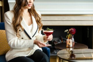 Woman in elegant jacket holding a cocktail with herbs in a stylish setting.