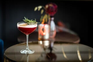 Cocktail mit rotem Getränk, Rosmarin und dekorativem Hintergrund auf einem Tisch.