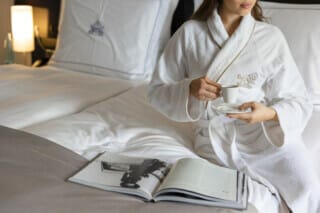 Hotel Atlantic Frau in Bademantel mit Tasse Kaffee, entspannt auf einem Bett mit Zeitschrift.
