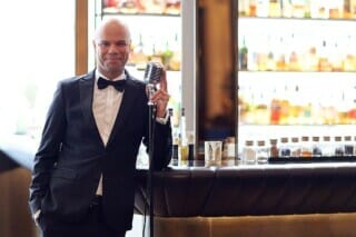 Man in a suit with a microphone at the bar, numerous bottles of spirits in the background.