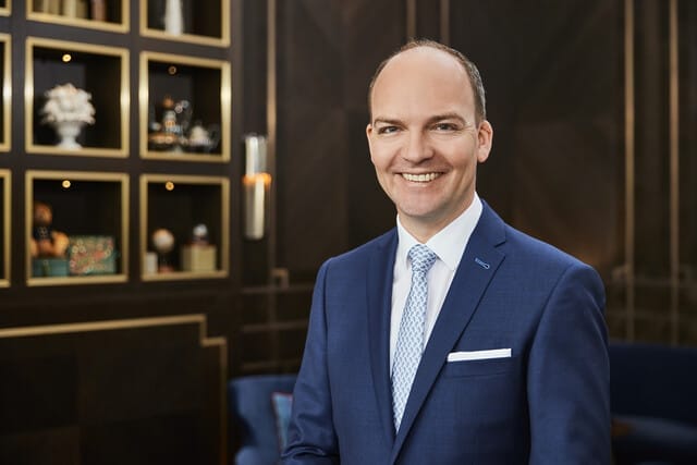 Professioneller Mann in Anzug lächelt vor elegantem Hintergrund mit Regalen.
