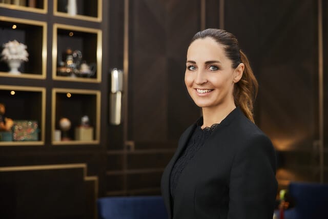 Professionelle Frau in schwarzem Blazer lächelt vor elegantem Hintergrund mit Regalen.