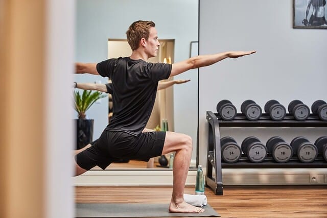 Mann in Sportkleidung übt Yoga in modernem Fitnessraum mit Gewichten im Hintergrund.